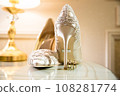 Elegant white bridal heels and wedding ring on a reflective surface, with a luxuriously blurred room backdrop. 108281774