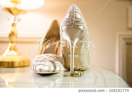 Elegant white bridal heels and wedding ring on a reflective surface, with a luxuriously blurred room backdrop. Elegant white bridal heels and wedding ring on a reflective surface, with a luxuriously blurred room backdrop. 108281774