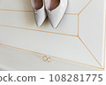 Elegant white bridal heels and wedding ring on a reflective surface, with a luxuriously blurred room backdrop. 108281775