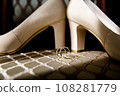 Elegant white bridal heels and wedding ring on a reflective surface, with a luxuriously blurred room backdrop. 108281779