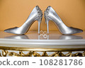 Elegant white bridal heels and wedding ring on a reflective surface, with a luxuriously blurred room backdrop. 108281786