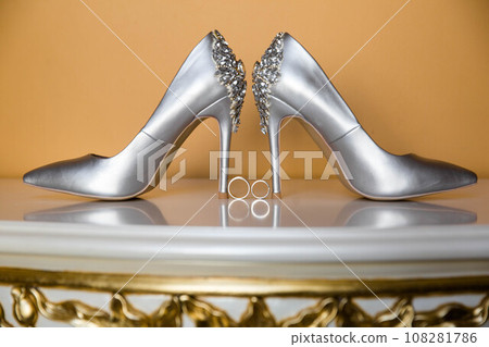 Elegant white bridal heels and wedding ring on a reflective surface, with a luxuriously blurred room backdrop. Elegant white bridal heels and wedding ring on a reflective surface, with a luxuriously blurred room backdrop. 108281786