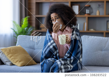 Sick cold woman sitting on sofa in living room, African American woman under blanket, has runny nose and high body temperature, sneezes and drinks hot drink with medicine. 108283522