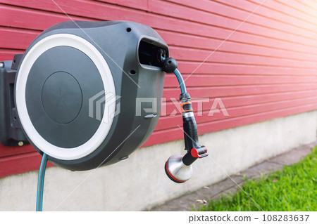 Wall Mounted Retractable Hose Reel. Modern garden lawn watering tip hose with sprinkler hanged at home wall. Lawn and plant tree smart irrigation water system equipment tool Wall Mounted Retractable Hose Reel. Modern garden lawn watering tip hose with sprinkler hanged at home wall. Lawn and plant tree smart irrigation water system equipment tool 108283637