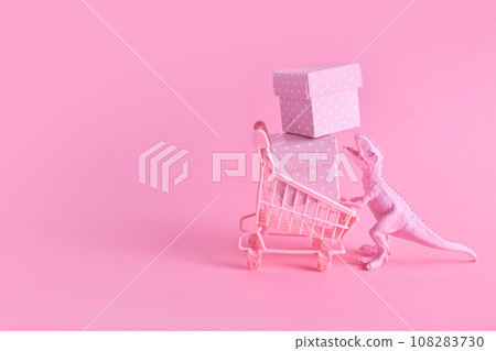 Pink Friday concept. Funny pink dinosaur toy with shopping cart full of present boxes on pink background 108283730