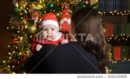 Cute baby in Santas hat and costume resting on the shoulder of mother near Christmas tree. Winter holidays and New Year concept Cute baby in Santas hat and costume resting on the shoulder of mother near Christmas tree. Winter holidays and New Year concept 108283809