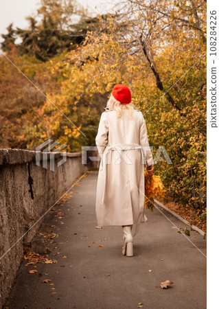 autumn woman in a red beret, a light coat and a red skirt, against the backdrop of an autumn park with yellow leaves in her hands 108284226