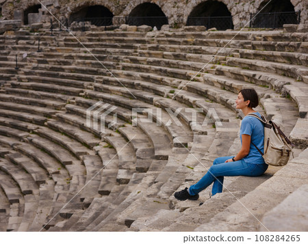 Woman tourist sitting in amphitheater enjoying solitude. travel to ancient city of Side, Antalya coast of Turkey in tourist low season. view of ancient theatre or Amphitheatre in Side, Turkey. Woman tourist sitting in amphitheater enjoying solitude. travel to ancient city of Side, Antalya coast of Turkey in tourist low season. view of ancient theatre or Amphitheatre in Side, Turkey. 108284265