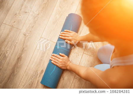 Woman hands rolled up yoga mat on gym floor in yoga fitness training room. Home workout woman close up hands rolling foam yoga gym mat. Woman barefoot home workout sportive healthy lifestyle concept 108284281