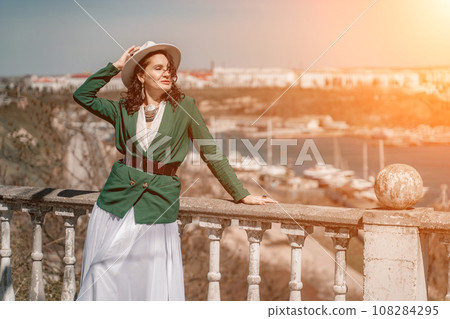 Woman walks around the city, lifestyle. Happy woman in a green jacket, white skirt and hat is sitting on a white fence with balusters overlooking the sea bay and the city. 108284295