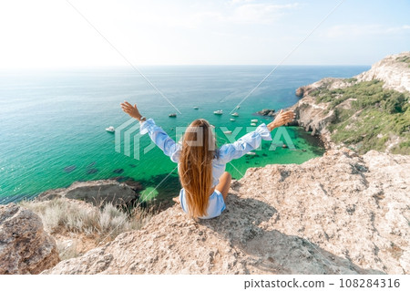 Woman travel sea. Happy woman in a beautiful location poses on a cliff high above the sea, with emerald waters and yachts in the background, while sharing her travel experiences 108284316