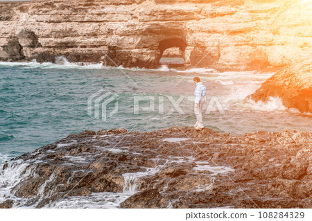 woman sea travel. A woman in a blue jacket stands on a rock above a cliff above the sea, looking at the stormy ocean. Girl traveler rests, thinks, dreams, enjoys nature woman sea travel. A woman in a blue jacket stands on a rock above a cliff above the sea, looking at the stormy ocean. Girl traveler rests, thinks, dreams, enjoys nature 108284329