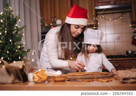 Happy little girl with her beautiful mom make gingerbread at home. Christmas decoration at kitchen. Fir tree with fairy lights Happy little girl with her beautiful mom make gingerbread at home. Christmas decoration at kitchen. Fir tree with fairy lights 108284383