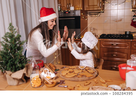 Mom in santas hat with her little doughter decorates gingerbread at home. Christmas and New Year traditions concept. Christmas bakery. Happy holidays 108284395
