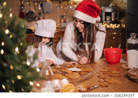 Mom with adorable little daughter spend time at kitchen in Christmas eve. Girls preparing sweets for holidays. Gingerbread and candy. Happy family 108284397