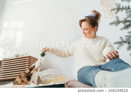 Cat pet is playing with glass bubble during young woman decorating Christmas tree with her cat in light modern Scandinavian interior. Winter festive holidays with domestic pet at home. Selective focus 108284628