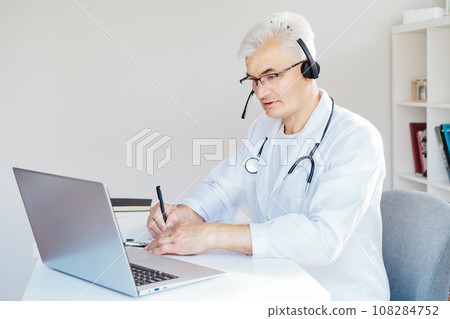 Middle aged male doctor using headset and laptop for online video call consulting of patient. Telemedicine concept for domestic health treatment. Online remote medical appointment. Medical technology. 108284752