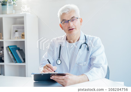 Confident mature middle aged man doctor medical nurse, male physician practitioner in white coat with stethoscope sitting at working place at hospital office and making notes during appointment 108284756