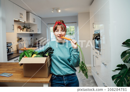 Smiling woman eating fresh carrot from her healthy food delivery box. Box with vegetables and phone with Veganuary app on the kitchen table. Start of a healthy life concept. Online home food delivery. 108284781