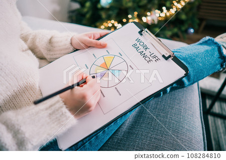 Woman drawing work-life balance wheel sitting on the sofa at home. Self-reflection and life planning for next new year. Personal Year review. Coaching tools. Finding Balance in Life. Selective focus. 108284810