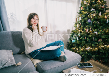 Young Woman with cup is hand writing New year's goals on note paper in new year day. New year resolutions list. Goals, resolutions, plans, action, checklist concept. New Year template, copy space Young Woman with cup is hand writing New year's goals on note paper in new year day. New year resolutions list. Goals, resolutions, plans, action, checklist concept. New Year template, copy space 108284862