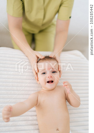 Vertical top view of adorable infant baby boy being manipulated by unrecognizable osteopathic manual therapist or physician. Closeup hands of female masseuse massage head of small newborn kid. 108285442