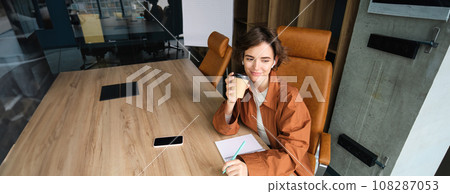 Portrait of employer working in office, woman entrepreneur in conference room, sitting at the table with documents, drinking coffee and smiling 108287053
