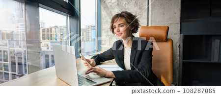 Portrait of businesswoman working on laptop, using mobile phone, sitting in an office, looking through her documents and workload, sharing coworking space 108287085