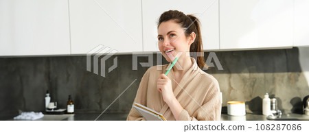 Portrait of beautiful modern woman, writing down grocery list, meal ideas in notebook, standing in the kitchen, wearing cosy bathrobe 108287086