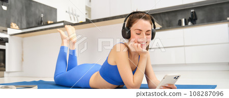 Portrait of fitness woman, listening yoga music in headphones, using her smartphone during workout at home, laying on blue sports mat 108287096