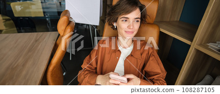 Portrait of woman working in an office, employee using smartphone, sitting in chair on her break and smiling 108287105