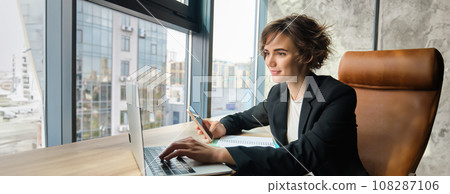 Portrait of businesswoman working on laptop, using mobile phone, sitting in an office, looking through her documents and workload, sharing coworking space 108287106