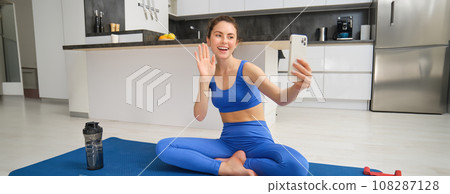 Vlogger does sports, records her workout training from home on smartphone camera, posing for selfie inside her house, sits on rubber yoga mat in blue leggings and sportsbra Vlogger does sports, records her workout training from home on smartphone camera, posing for selfie inside her house, sits on rubber yoga mat in blue leggings and sportsbra 108287128