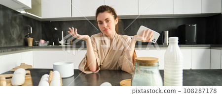 Attractive young cheerful girl baking at the kitchen, making dough, holding recipe book, having ideas Attractive young cheerful girl baking at the kitchen, making dough, holding recipe book, having ideas 108287178