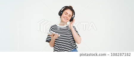 Young woman with smartphone listening to music, dancing to her favourite song in headphones, posing against white background 108287182