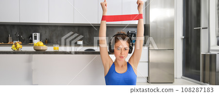 Workout and fitness concept. Portrait of beautiful, focused woman with arms raised above head, stretching with rubber resistance band, doing sport exercises, aerobics at home, wearing headphones 108287183