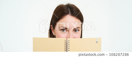 Young woman holds notebook daily planner next to her face, writing down homework, making notes, looking thoughtful, standing over white background 108287286