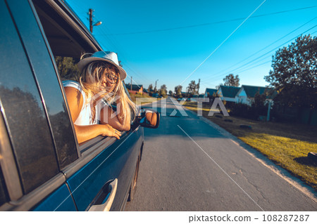 Leaning out the car window while driving, a woman smiles and looks into the camera. The wind beautifully develops the woman's hair in the car window. The woman enjoys the feeling of freedom. 108287287