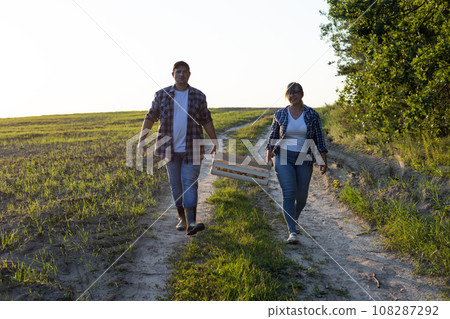 A couple of farmers carry a box of potatoes across the field against the backdrop of a beautiful sunset A man and a woman together carry a heavy box of vegetables harvested and grown in their fields. 108287292