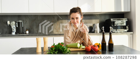 Healthy lifestyle. Young woman in bathrobe preparing food, chopping vegetables, cooking dinner on kitchen counter, standing over white background Healthy lifestyle. Young woman in bathrobe preparing food, chopping vegetables, cooking dinner on kitchen counter, standing over white background 108287298