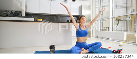 Portrait of girl doing yoga, fitness training, raising hands up and breathing, mindful practices, sitting on rubber mat in living room, exercising Portrait of girl doing yoga, fitness training, raising hands up and breathing, mindful practices, sitting on rubber mat in living room, exercising 108287344