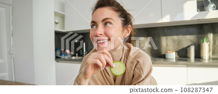 Portrait of happy, smiling young woman in the kitchen, cooking, chopping zucchini, holding vegetables and looking happy, preparing vegan food meal at home Portrait of happy, smiling young woman in the kitchen, cooking, chopping zucchini, holding vegetables and looking happy, preparing vegan food meal at home 108287347