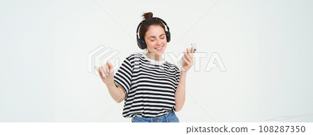 Girl with smartphone dancing, listening to music in wireless headphones, using mobile phone streaming app to enjoy favourite songs, isolated over white background 108287350