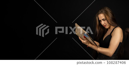 Halloween. Portrait. A beautiful woman with black makeup holds an ancient spell book in her hands on a black background. The witch is studying an old book holding it in her hands. Copy space. 108287406