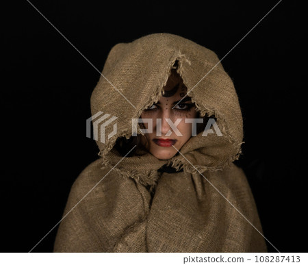 In a cape with a hood, a beautiful female witch stands in the semi-darkness with runic symbols on her face. Halloween. Halloween black witch portrait. Halloween holiday concept. 108287413