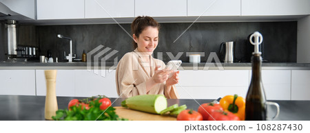 Portrait of woman in bathrobe sitting in kitchen with smartphone, cooking dinner, watching recipe on social media, video tutorial how to prepare meal Portrait of woman in bathrobe sitting in kitchen with smartphone, cooking dinner, watching recipe on social media, video tutorial how to prepare meal 108287430