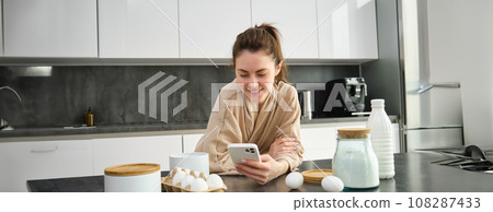Attractive young cheerful girl baking at the kitchen, making dough, holding recipe book, having ideas Attractive young cheerful girl baking at the kitchen, making dough, holding recipe book, having ideas 108287433