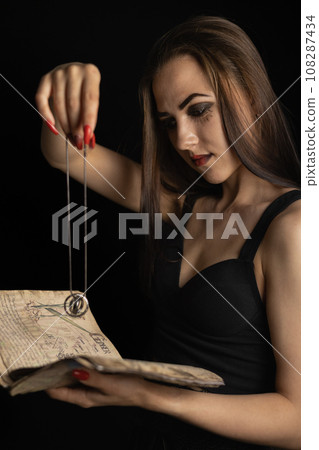On Halloween, the witch performs a ritual with a book and a talisman. A beautiful female witch reads fortunes from an ancient book, holding a talisman in her hand. Halloween divination concept. 108287434