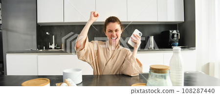 Attractive young cheerful girl baking at the kitchen, making dough, holding recipe book, having ideas Attractive young cheerful girl baking at the kitchen, making dough, holding recipe book, having ideas 108287440