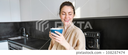 Portrait of carefree smiling woman drinking coffee, standing in kitchen with delighted, pleased face expression, waking up in the morning and having cappuccino Portrait of carefree smiling woman drinking coffee, standing in kitchen with delighted, pleased face expression, waking up in the morning and having cappuccino 108287474